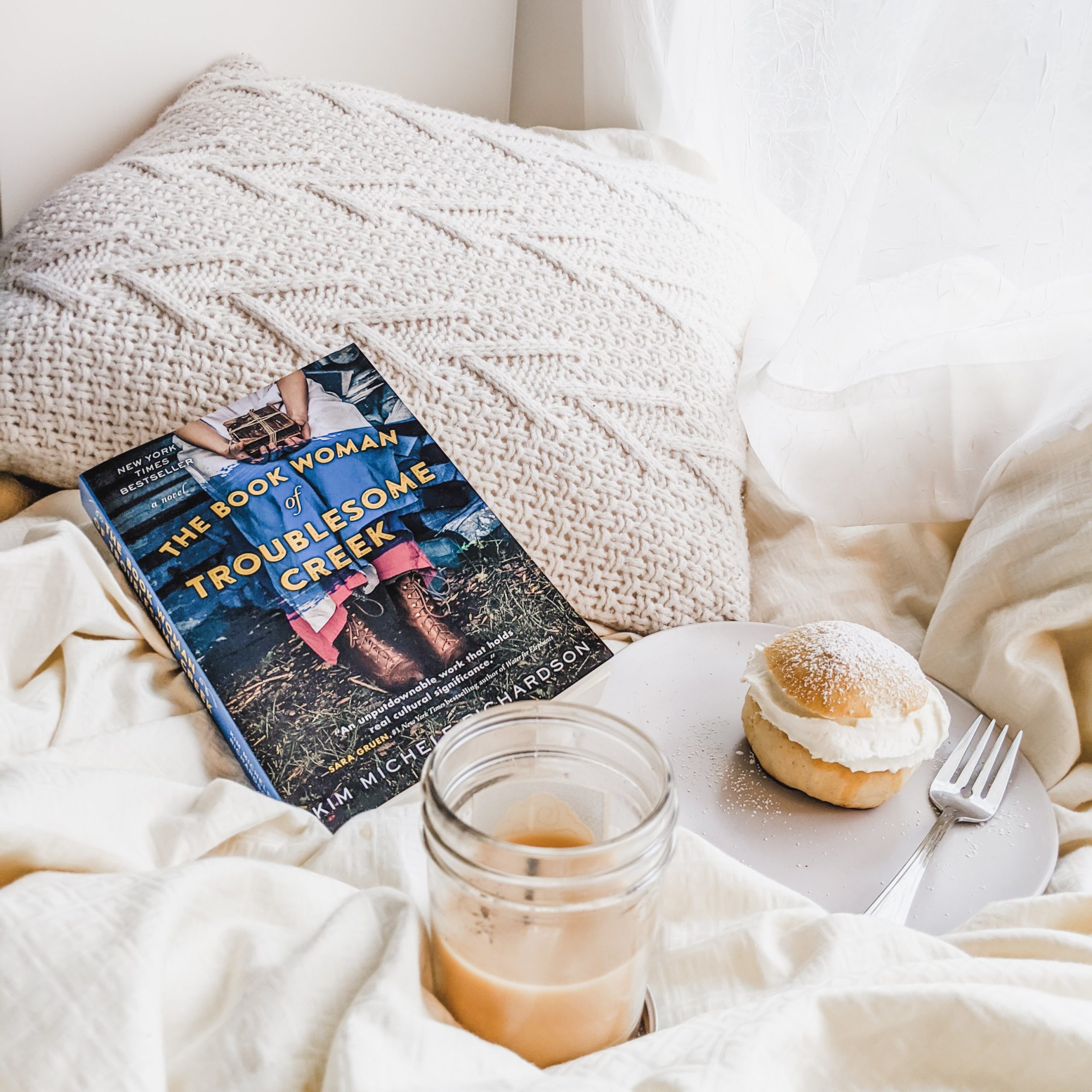 The Book Woman of Troublesome Creek book sitting on white covers by a dessert and candle