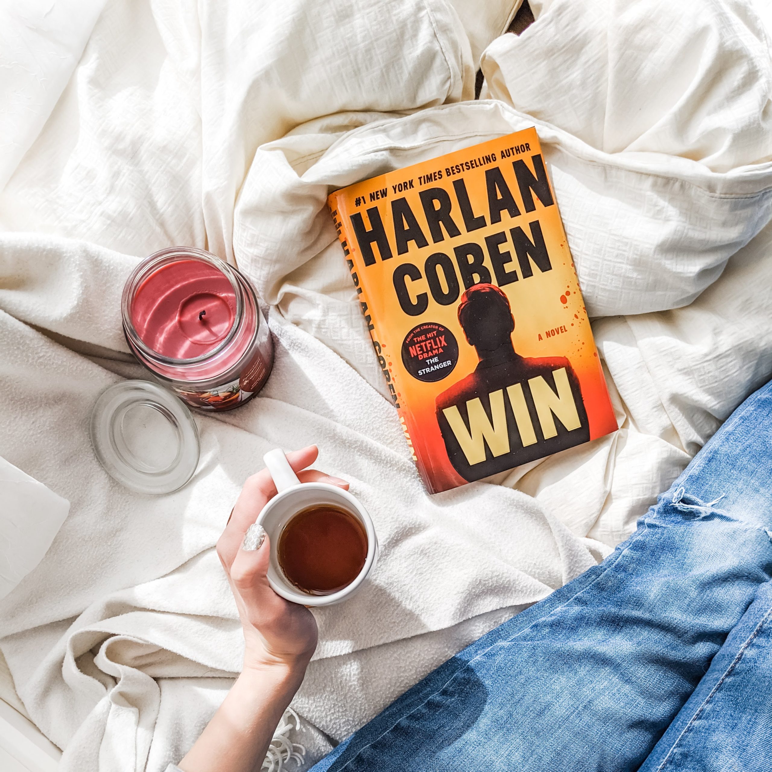 Win by Harlan Coben book on white blankets. Girl holds mug of coffee.