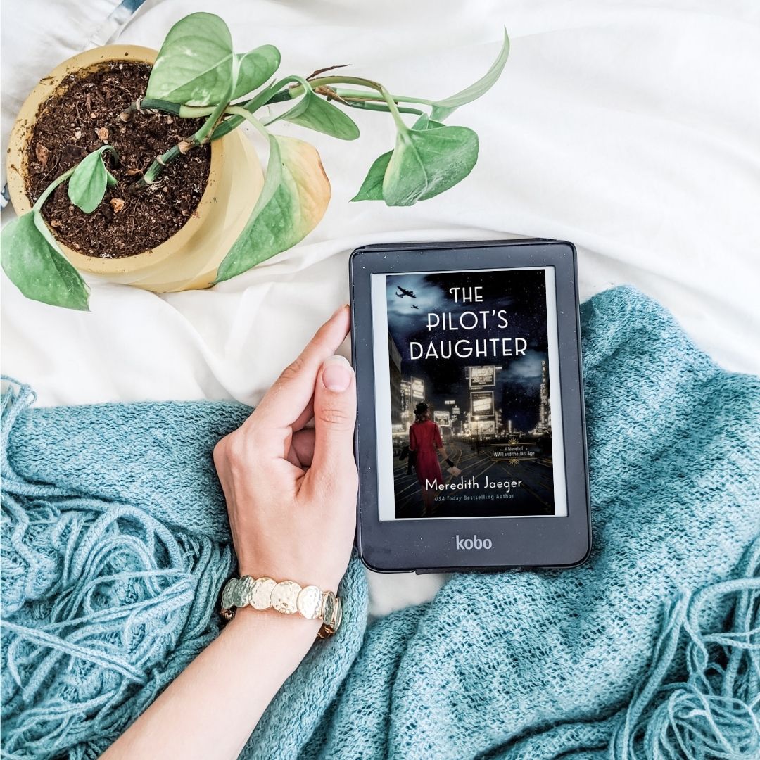 The Pilot's Daughter. Kobo reader set against white background and blue blanket with potted plant.