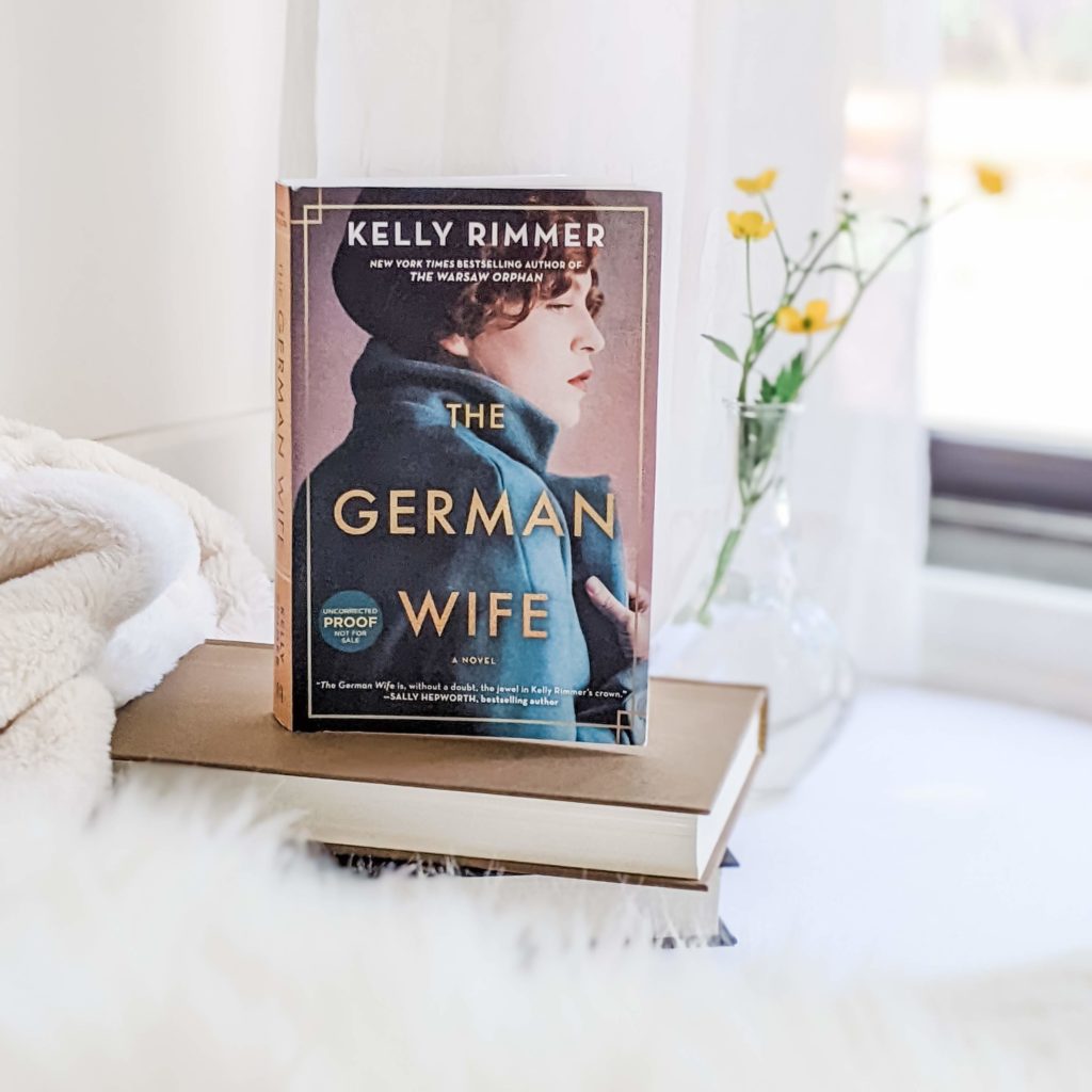 The German Wife by Kelly Rimmer, photo of book cover set against white background with yellow flowers. By The 52 Book Club